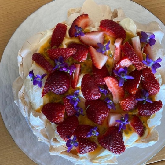 Aerial photo of a Pavloa on a white plate, edges of the off-white meringue can be seen under a bed of cut red strawberries, passion fruit seeds and vibrant purple violets, the shape of the pavlova is round
